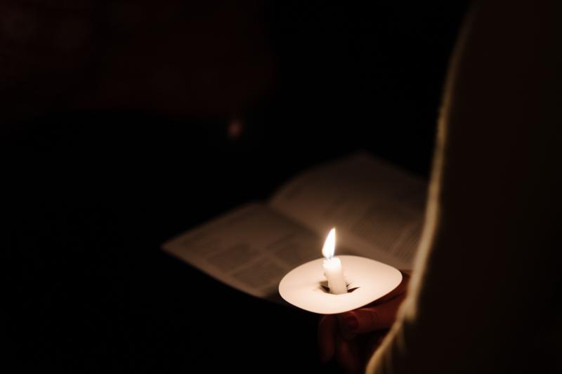 Taper candle casting glow on person and paper program at a prayer vigil.